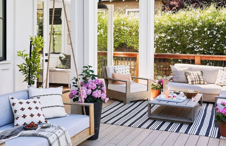 Stylish patio with light wood furniture, pastel cushions, potted flowers, and a black-striped rug