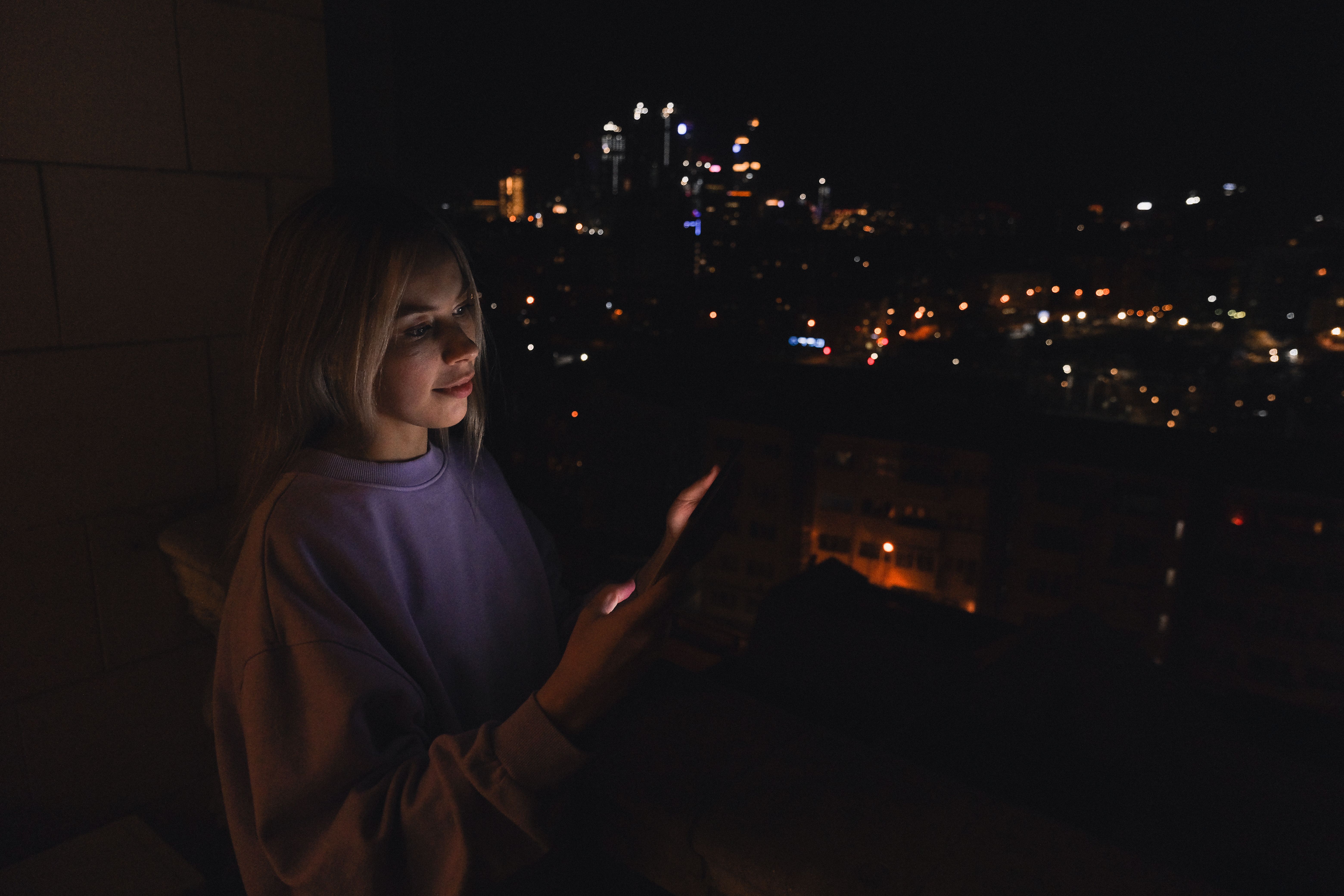 Woman on a balcony at night illuminated by her smartphone screen, illustrating late-night mobile gaming