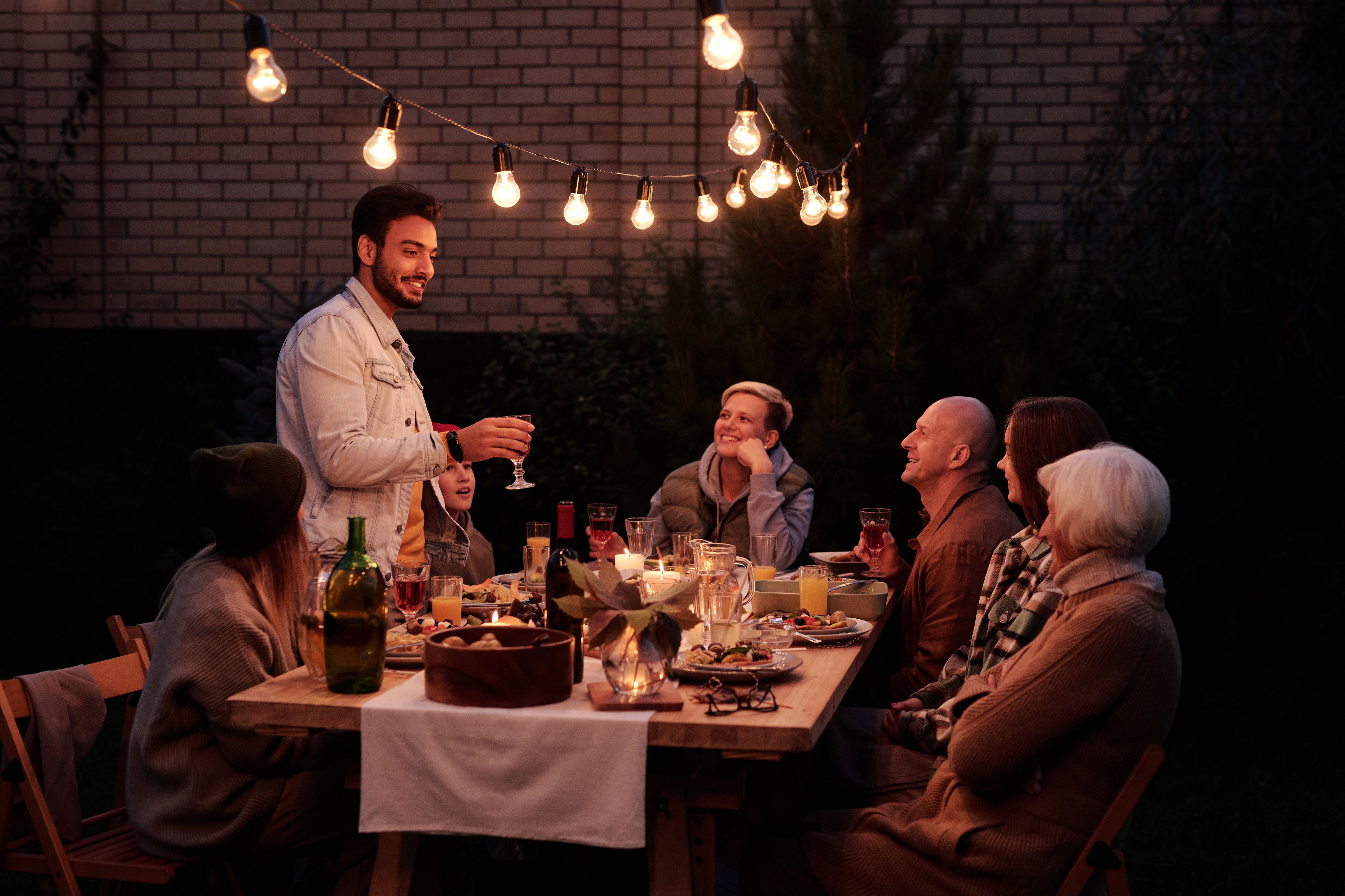 Friends sharing food and conversation at an outdoor dinner party under warm string lights.